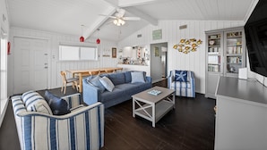 Ein Wohnzimmer mit einer Couch, einem Couchtisch und gestreiften Stühlen. Es gibt einen Essbereich mit Tisch und Stühlen. Im Hintergrund ist eine Küche zu sehen.