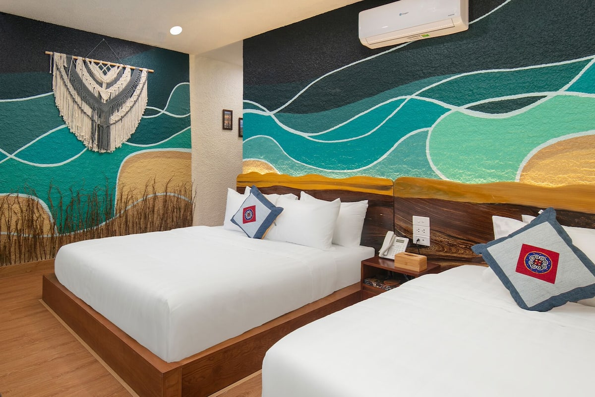 A hotel room with two beds, a mural on the wall, and a small table with a fruit bowl.