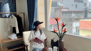 Hotellihuone, jossa on suuri ikkuna, tuoli, pöytä, jossa on maljakko täynnä kukkia, sekä pyyhe- ja vaateteline.