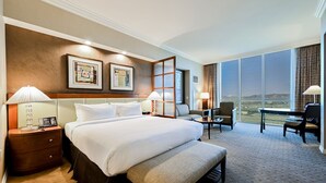 A hotel room with a large bed, two bedside tables with lamps, a desk with a chair, and a large window offering a view. - SVS: Penthouse 2BD/3BA | Mountain View Balcony | Full Kitchen | MGM (Las Vegas)