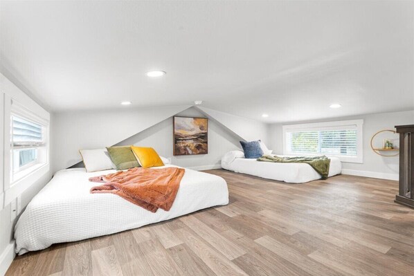 Un dormitorio con techo inclinado, una cama con cubrecama blanco y manta naranja, un sofá blanco con cojines, un cuadro en la pared y una ventana con persianas.