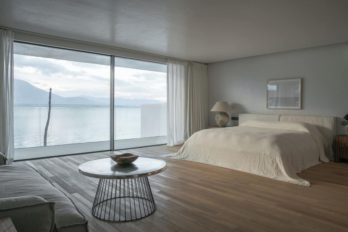 Une chambre d’hôtel moderne avec une grande fenêtre, une table ronde sur laquelle se trouve un bol, un lit avec du linge de lit blanc et une lampe sur une table de chevet.