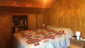 A cozy bedroom with a wooden ceiling, a bed with a patterned blanket, a nightstand with a lamp, and a bookshelf filled with books. - Chalet Ours Blanc ORS Nature & Quiet 6 Pers (Chatel)