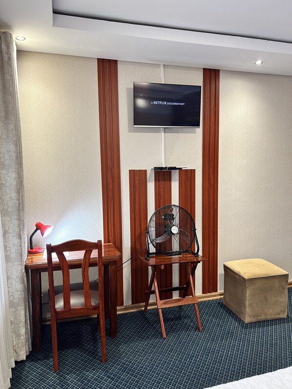 Une chambre d’hôtel avec un bureau en bois, une chaise, un petit pouf, un ventilateur sur pied, une lampe de bureau et un téléviseur à écran plat fixé au mur.