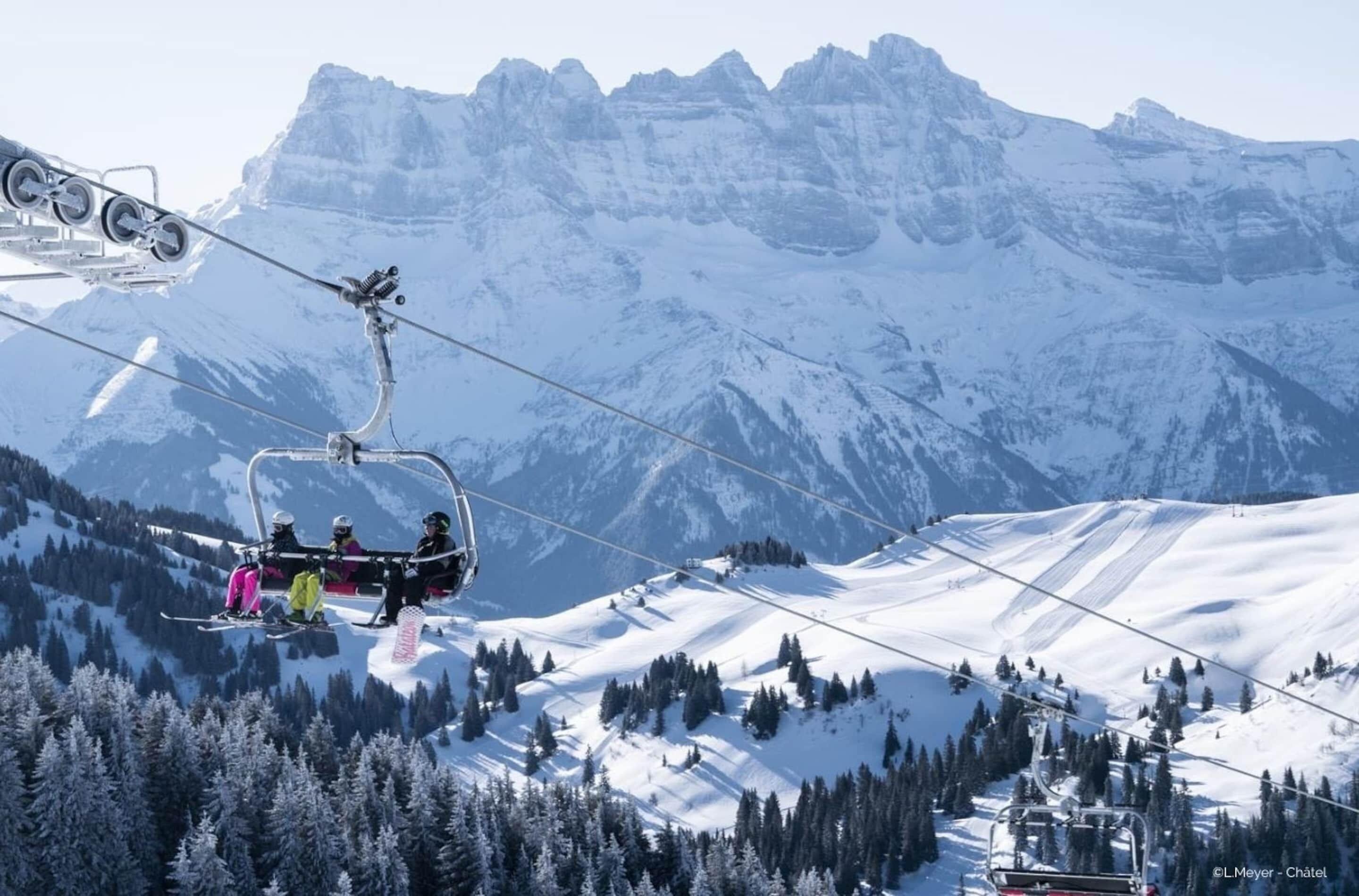 A ski lift with skiers in colorful gear, surrounded by snow-covered mountains and trees.