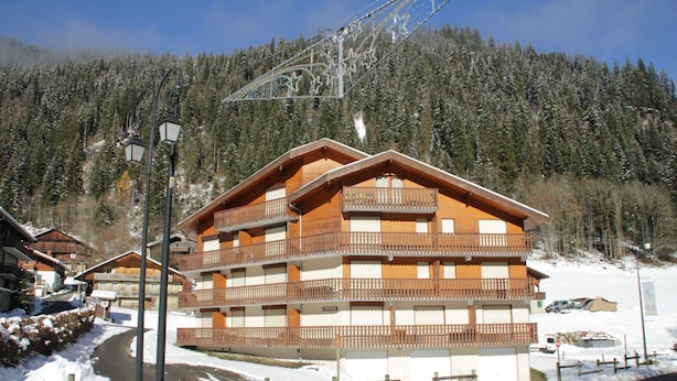 A multi-story wooden building with balconies, surrounded by snow-covered trees and a clear sky. - Orchidee OD5 Nature & Telecabine 4 Pers (Chatel)