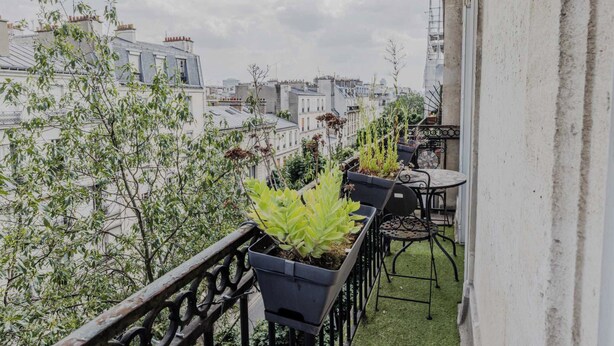 Ein Balkon mit Blumentöpfen und einem Tisch, mit Blick auf eine Stadtlandschaft aus Gebäuden und einem bewölkten Himmel.