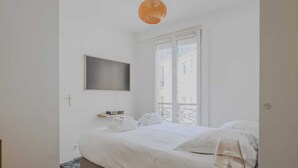 Ein Hotelzimmer mit Bett, Nachttisch mit Lampe, Fenster mit Blick auf Gebäude und einem an der Wand montierten Fernseher.
