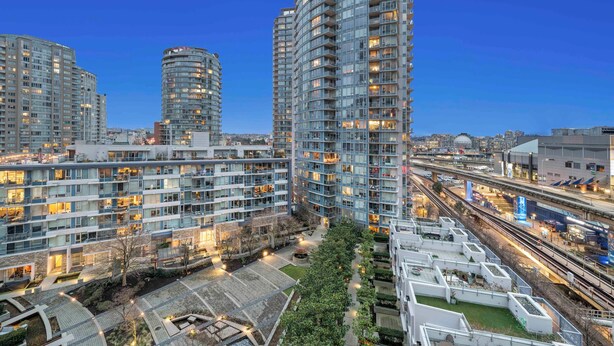 Un edificio residencial de gran altura con varios balcones, un jardín en la azotea y vistas a una concurrida calle urbana con vías de tren.