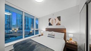 Una habitación de hotel moderna con una cama grande, una mesita de noche, una lámpara y vistas a un paisaje urbano a través de amplios ventanales.