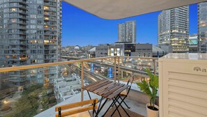 Un balcón con mesa y banco de madera, una planta en maceta y vistas de un paisaje urbano con edificios modernos y una autopista.