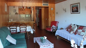 Una sala de estar acogedora con un sofá verde, un sillón rojo y una cama con ropa de cama estampada. También se ve un comedor con muebles de madera y una cocina al fondo.