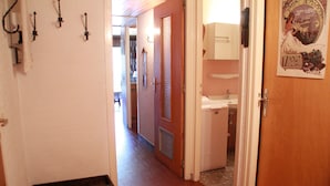 Un pasillo estrecho con una puerta de madera a la derecha, una pared blanca con un cartel, y una pequeña área de cocina.