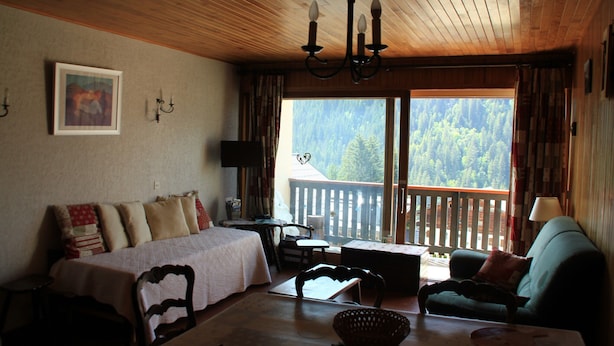Una habitación acogedora con cama, una mesa de comedor, una silla, un sofá y vistas a un bosque a través de un ventanal grande.