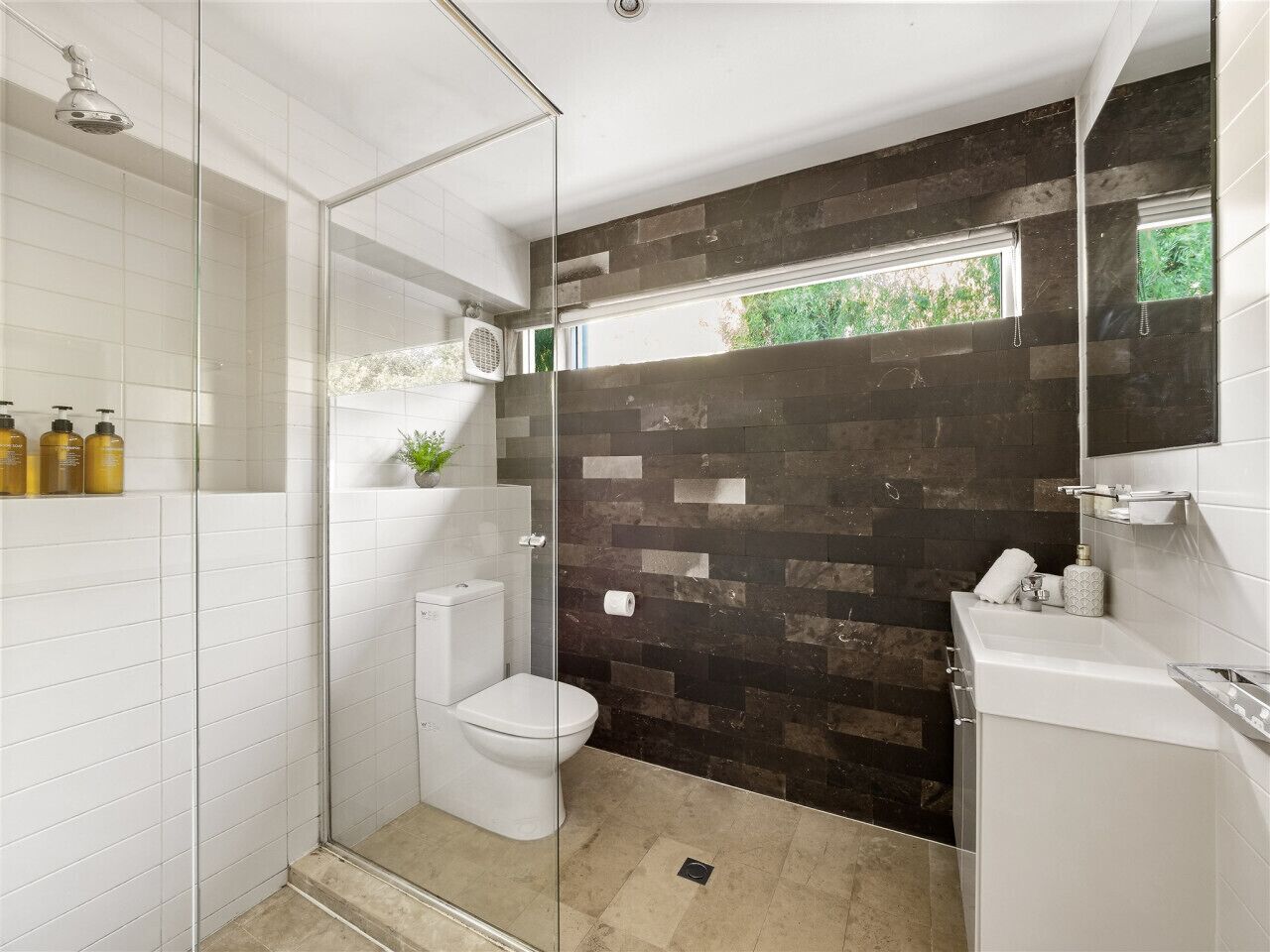 Une salle de bain moderne avec une douche à l’italienne en verre, des toilettes blanches et un mur recouvert de carreaux foncés.