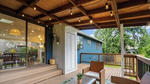 A covered deck with wicker furniture, a glass door leading to a patio, and a blue house in the background. - Spacious Modern Retreat w Game Room and Yard (Lynnwood)