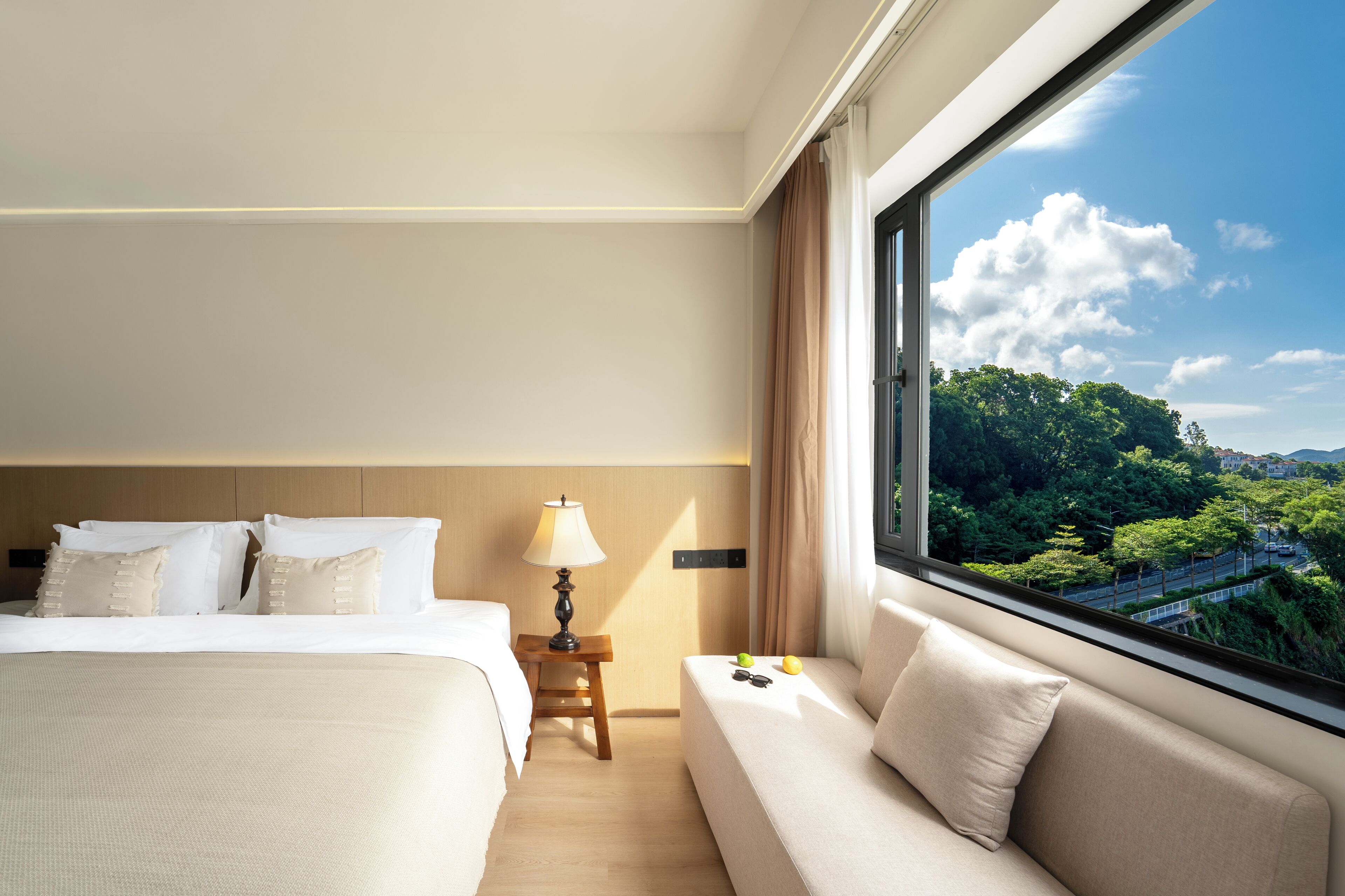 Habitación de hotel con cama, sofá, mesita de noche con lámpara y ventana con vistas a zonas verdes.