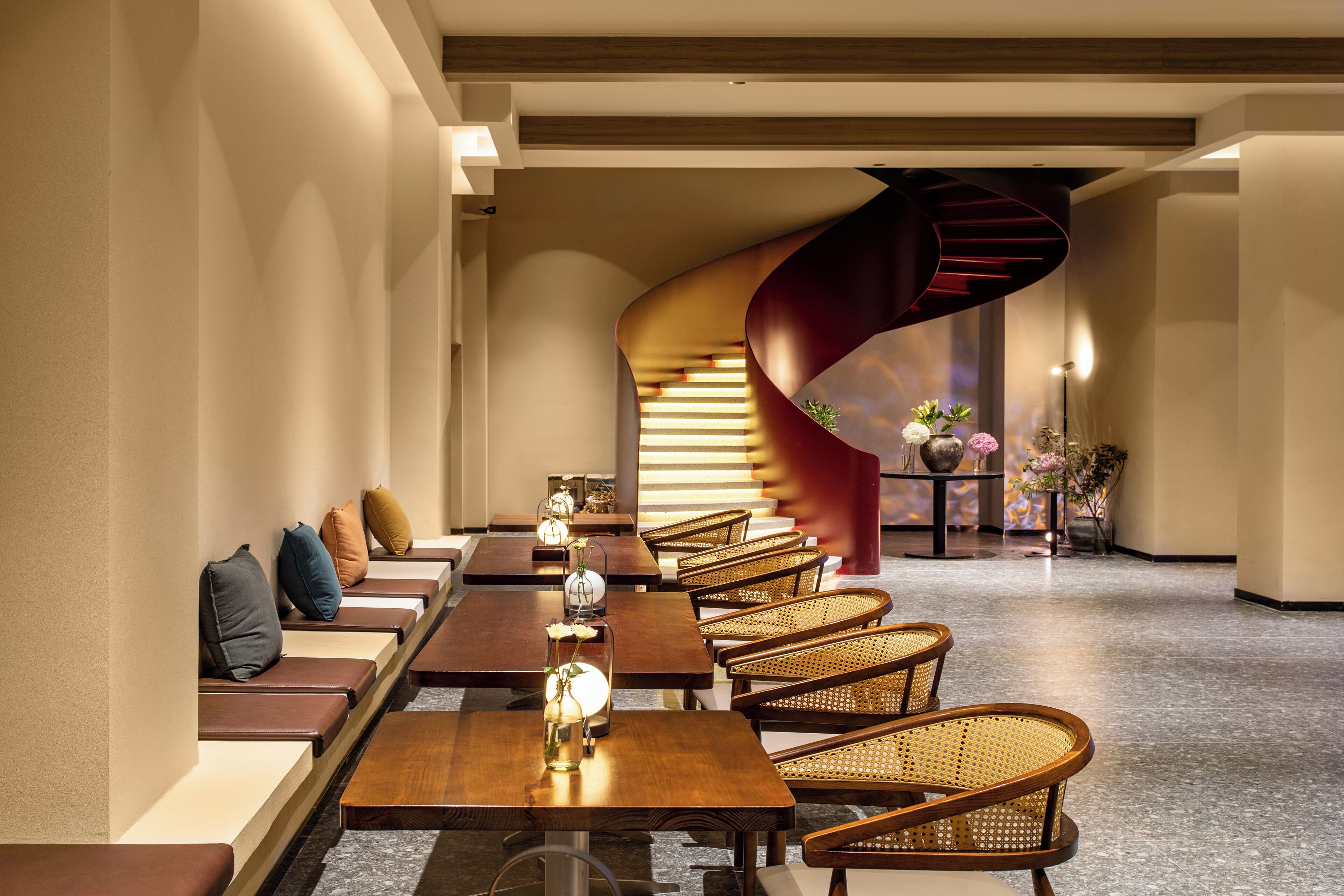 Un moderno vestíbulo de hotel con largas mesas de madera y sillas de mimbre, una escalera en espiral y un florero sobre una mesita.