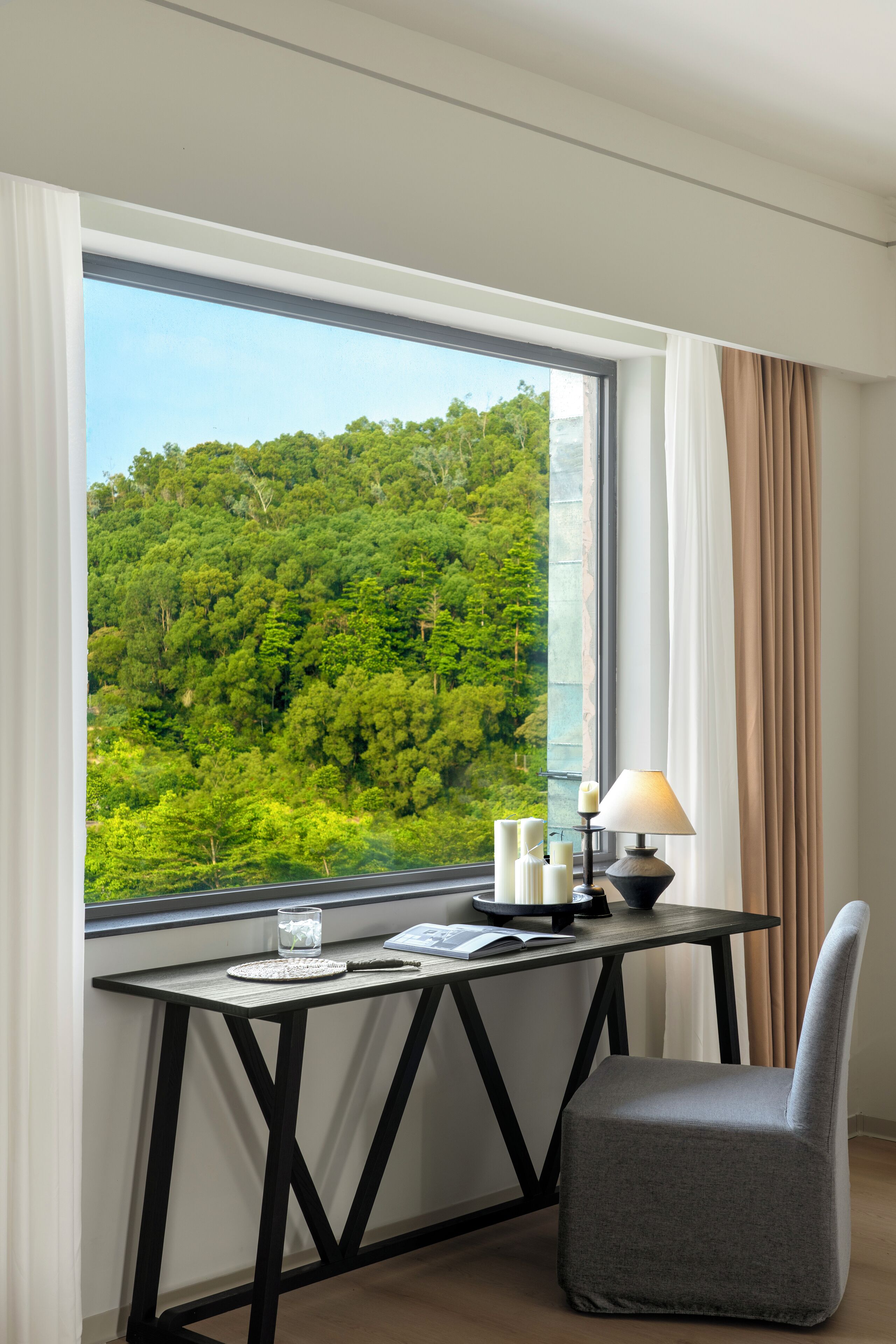 A hotel room with a desk and chair, a window with a view of a forest, and a lamp on the desk.