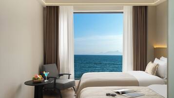 A hotel room with a bed, a chair, a small table with a drink and fruit, and a view of the sea.