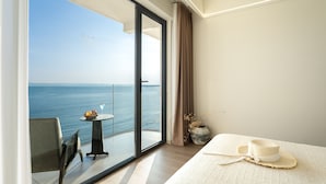A modern hotel room with a large balcony, a dining table with fruit and a drink, and a bed with a hat and a vase.