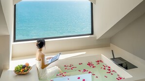 A room with a large window overlooking the sea, a bathtub with rose petals, and a fruit bowl on a bench.