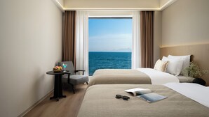 A hotel room with a large window, a bed, a chair, a small table with a fruit bowl and a drink, and a nightstand with a vase of flowers.