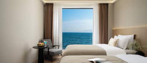 A hotel room with a large window, a bed, a chair, a small table with a fruit bowl and a drink, and a nightstand with a vase of flowers.