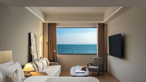A hotel room with a large window offering a sea view, a bed, a sofa, a chair, a small table with a bowl of fruit, and a flat-screen TV mounted on the wall.