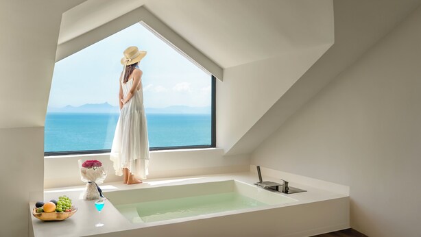A woman in a white dress standing by a bathtub with a view of the sea.