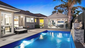 A modern outdoor pool area with a clear blue pool, a covered seating area with cushions, and a stone wall with a statue. - Resort Experience: Pool, BBQ, Games with lots of space and fun in Pakenham (Pakenham)