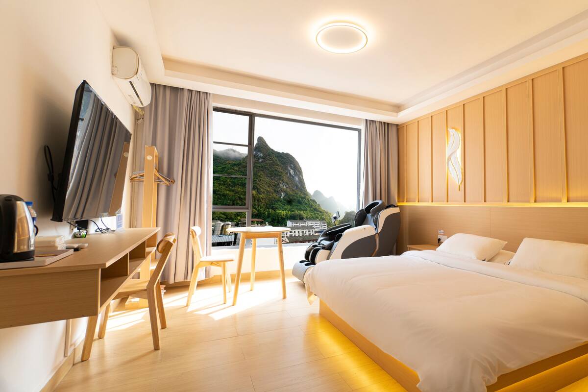 Een hotelkamer met een bed, bureau, stoel en uitzicht op bergen door grote ramen.