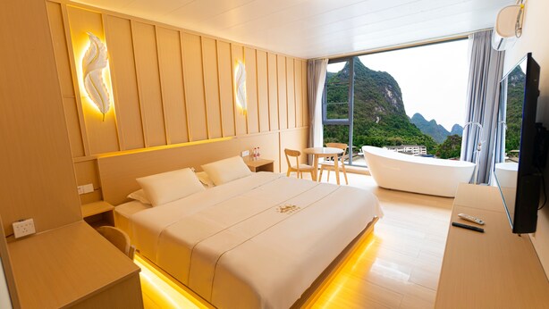 A modern hotel room with a large bed, a small table, a chair, and a view of mountains through the window. - Shanxun Snow Mountain Villa Hotel (Guilin)