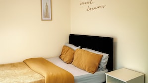 Ein ordentlich bezogenes Bett mit schwarzem Kopfteil, zwei Kissen und goldener Tagesdecke. Ein kleiner Nachttisch ist rechts sichtbar.