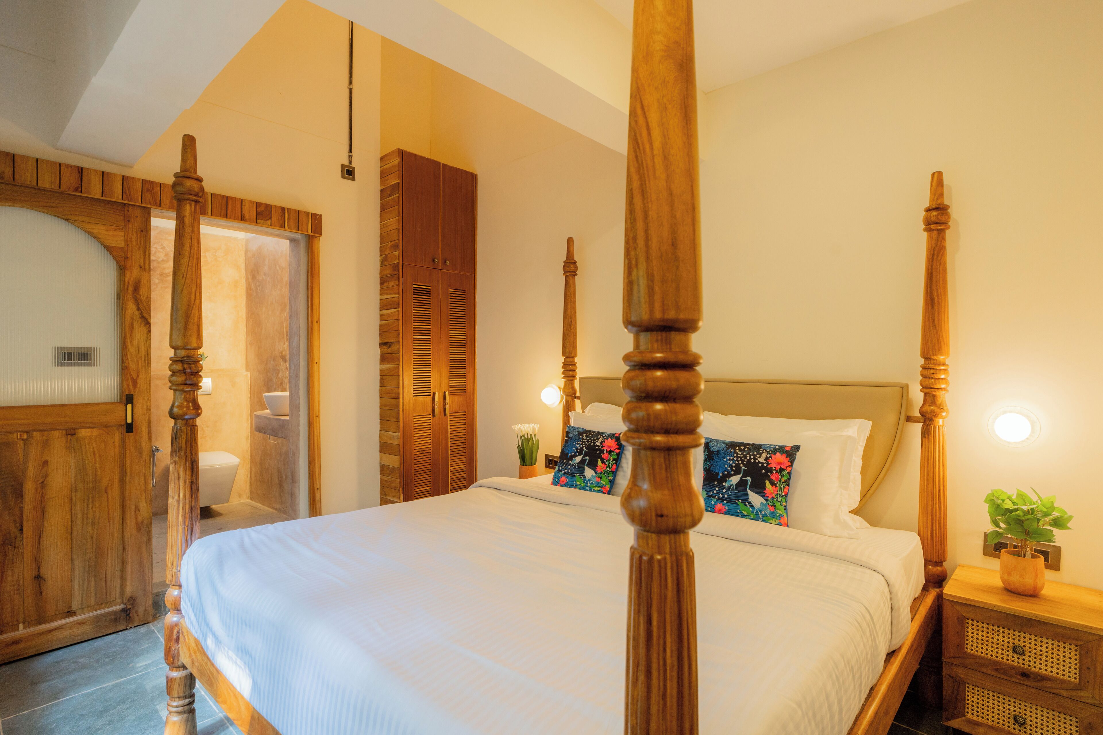 A four-poster bed with wooden posts, a white bedspread, and floral pillows. There is a wooden wardrobe and a nightstand with a plant.