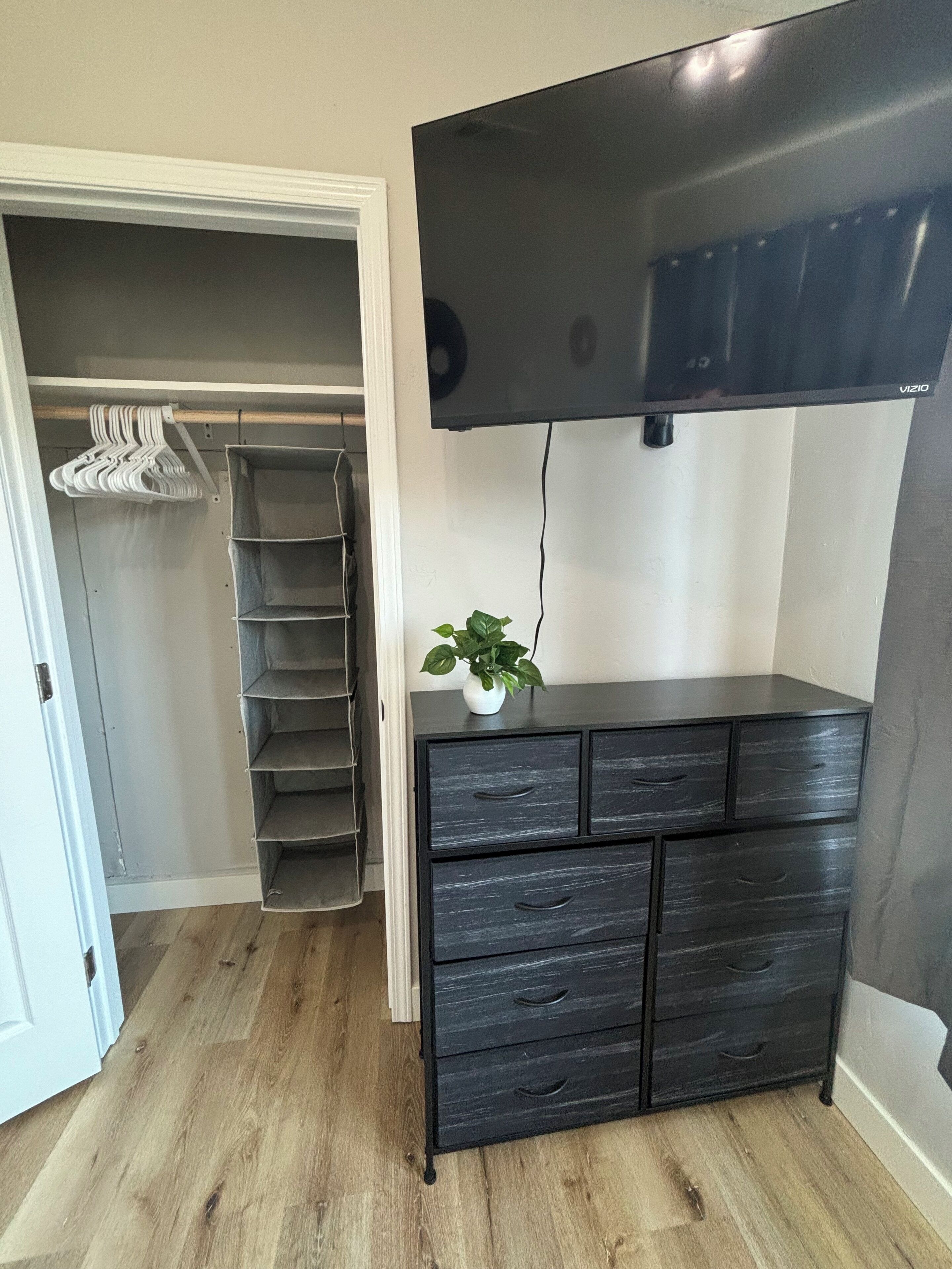 A room with a wooden floor, a black dresser with drawers, a flat-screen TV mounted on the wall, and a white door leading to a closet with hanging clothes.