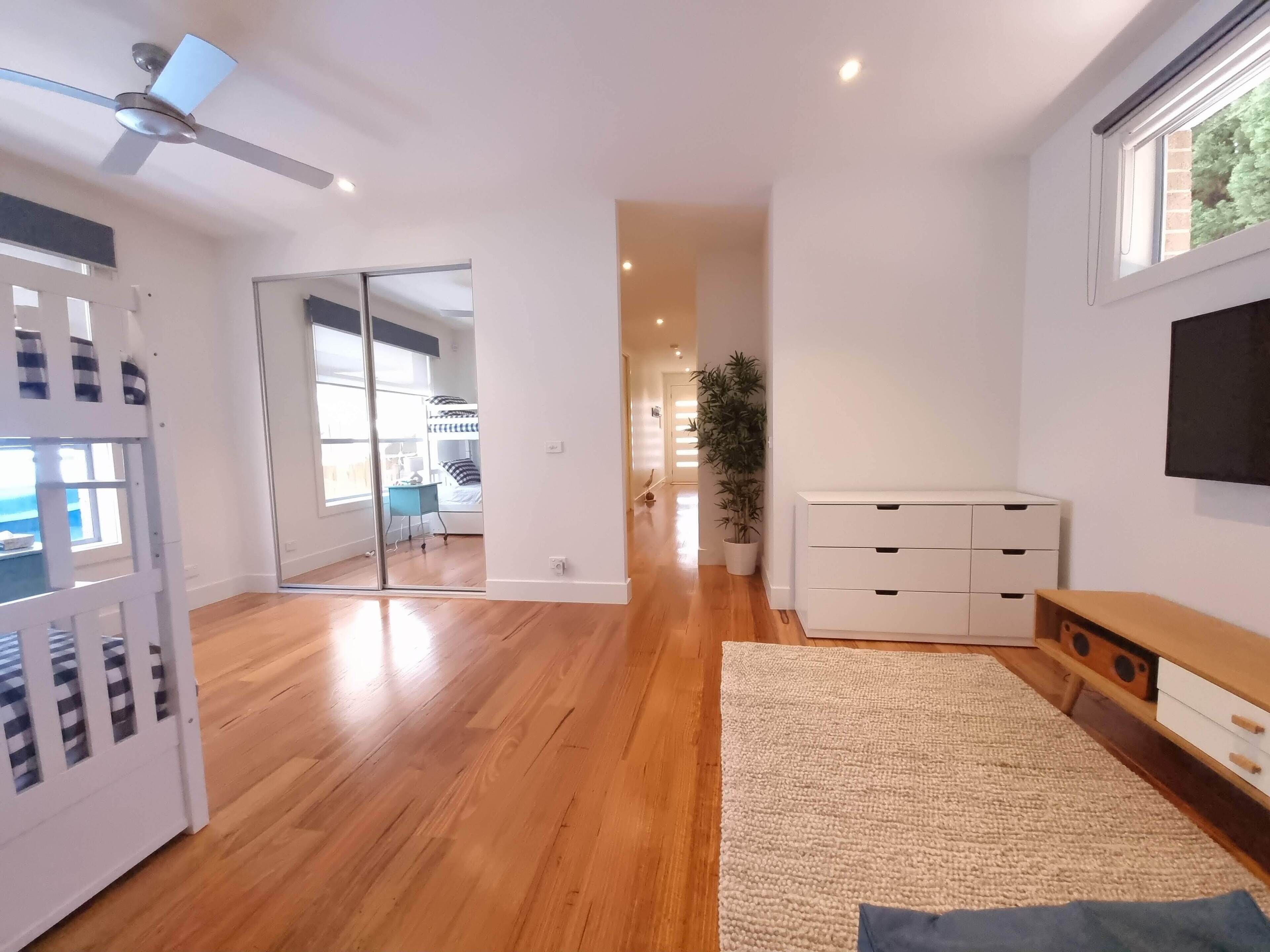 A spacious room with wooden flooring, a ceiling fan, a large window, a white cabinet, and a TV mounted on the wall.
