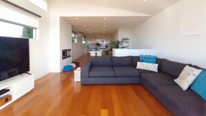 Un salon moderne avec un grand canapé d’angle, un téléviseur à écran plat, un plancher en bois et une cuisine en arrière-plan.
