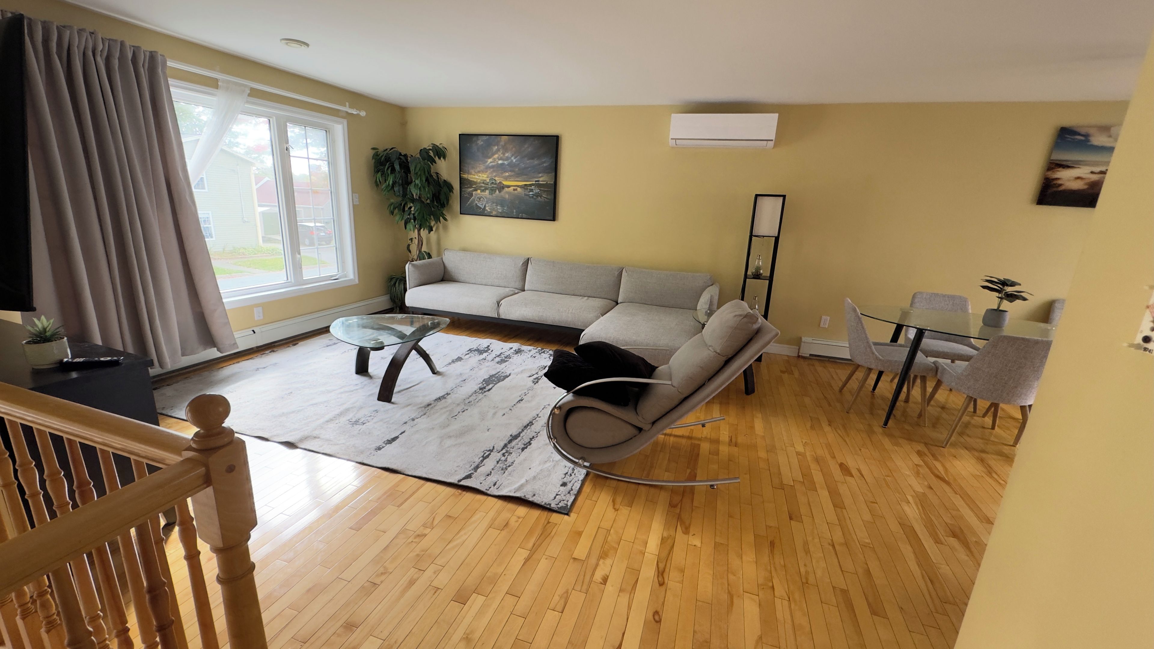 Una sala de estar con un sofá seccional, una mesa de centro de vidrio, una zona de comedor con una mesa pequeña y sillas, y un ventanal con cortinas.