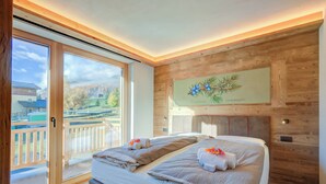 A bedroom with a large sliding glass door leading to a balcony, a bed with neatly folded towels, and a wooden wall with a framed botanical illustration. - New Ginepro Ski 10 m, Livigno, Italy (Livigno)