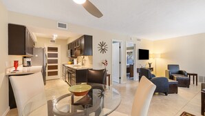 A modern living room with a glass dining table, a ceiling fan, and a kitchen in the background.