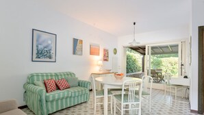 Een gezellige woonkamer met een groen gestreept bankje, een eettafel met stoelen, en uitzicht op groen buiten.