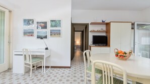 Een compacte kamer met een bureau, stoel en een eettafel met stoelen. Er is een televisie op een plank en een mand met fruit op de tafel.