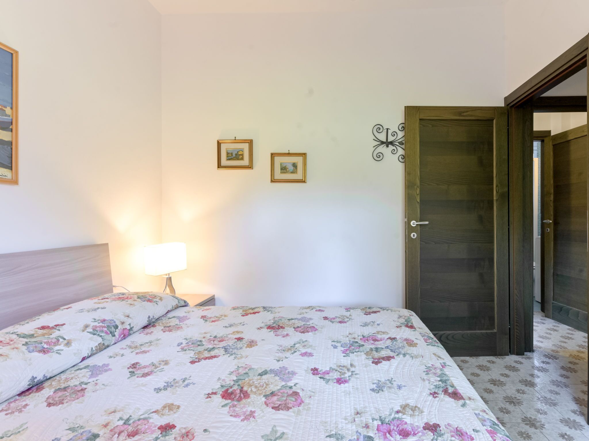 A bedroom with a bed, a nightstand, a lamp, two framed pictures, a decorative wall piece, and a wooden door.