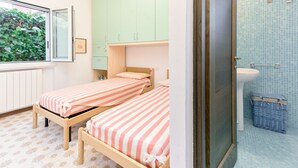 Een slaapkamer met twee eenpersoonsbedden, een raam met uitzicht op groen, een badkamer met een wastafel en blauwe betegelde muren.