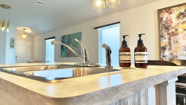 A kitchen with a stainless steel sink, a modern faucet, and pendant lights. There are two bottles of hand soap and dish soap on the counter.