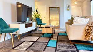 A modern living room with a flat-screen TV, teal chairs, a wooden coffee table, and a patterned rug.