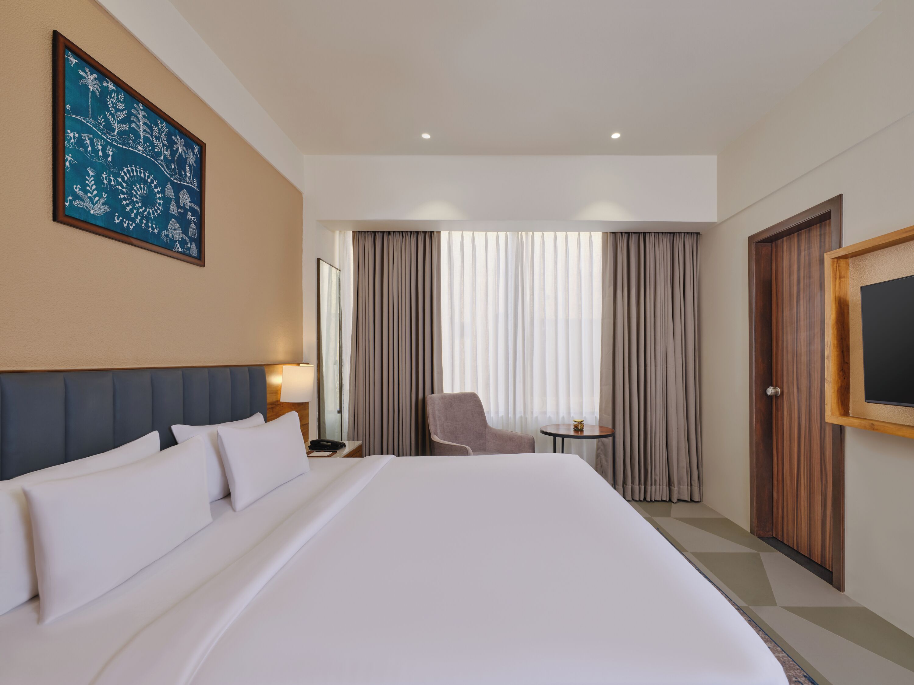 A hotel room with a large bed, a chair, a small table, a mirror, and a painting on the wall.
