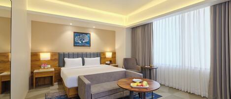 A hotel room with a large bed, a grey sofa, a small round table with fruit, a painting on the wall, and a large window with curtains.