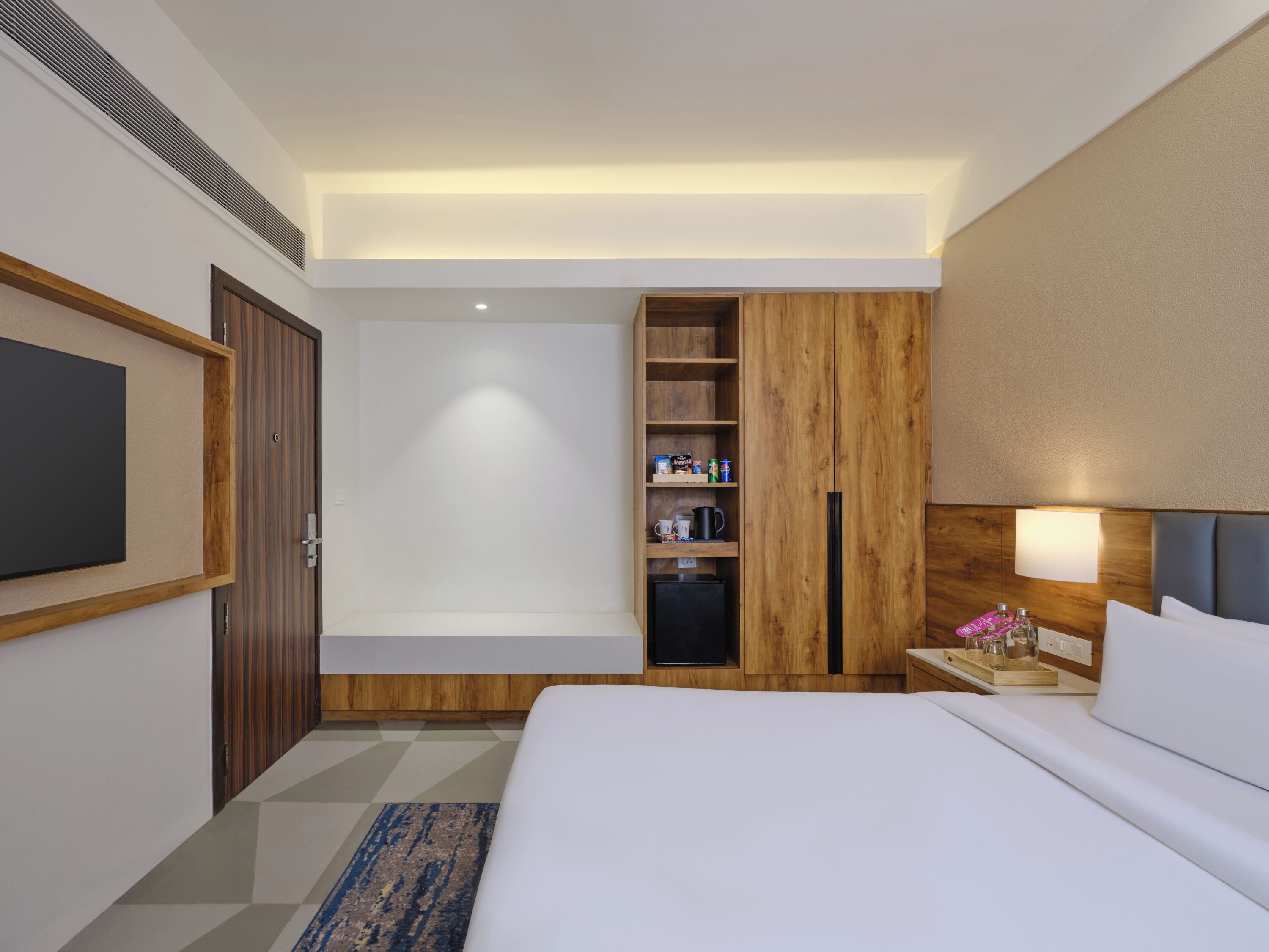 Habitación de hotel con cama, televisión, armario, repisa con artículos de aseo y mesita de noche con lámpara.